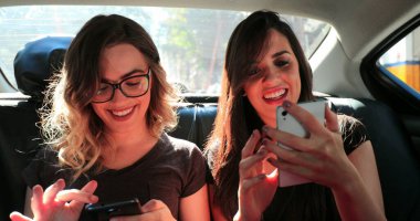 Two girls in the back seat of a car checking cellphone together. Female friends browsing on their smartphone while riding cab