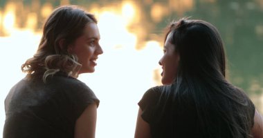 Friends talking by a lake. Girlfriends exchanging ideas and in conversation speaking to one another outdoors in nature