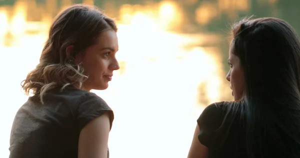 Friends talking by a lake. Girlfriends exchanging ideas and in conversation speaking to one another outdoors in nature