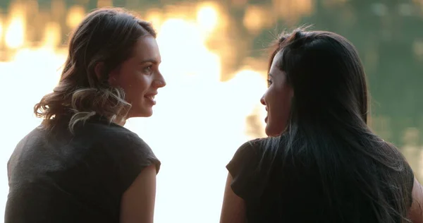 Friends talking by a lake. Girlfriends exchanging ideas and in conversation speaking to one another outdoors in nature