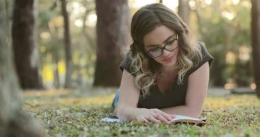 Smart intelligent girl wearing glasses lying on grass outside at the park reading book studying and turning page
