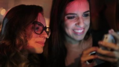 Woman showing media to her passenger friend. Girl shows something to girlfriend seated in the back of a car at night starring at smartphone screen