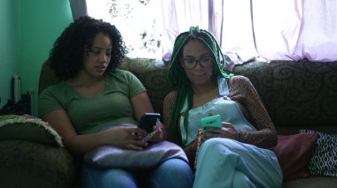 Two young black latina women looking at their phones sitting on sofa at home