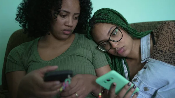 Two black latina women looking at phones sitting on sofa couch at home
