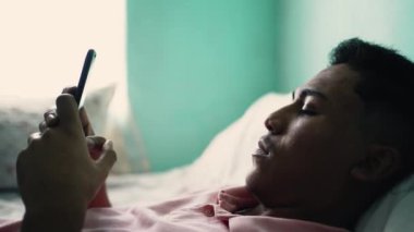 A hispanic man looking at cellphone device lying in bed staring at phone screen with serious expression