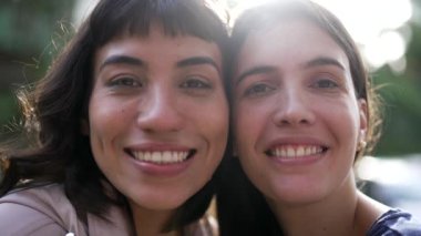Two female friends together looking at camera. Portrait of diverse girls outside with sun flare