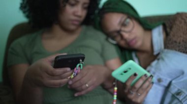 Two black latina women looking at phones sitting on sofa couch at home