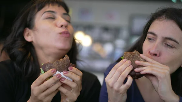 Two happy female friends eating burgers at the same time. Young women taking a bite of cheeseburgers at restaurant