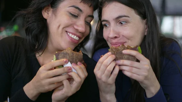 Two happy female friends eating burgers at the same time. Young women taking a bite of cheeseburgers at restaurant