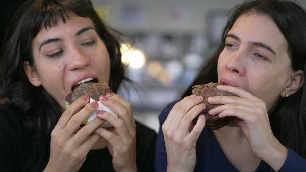 Two happy female friends eating burgers at the same time. Young women taking a bite of cheeseburgers at restaurant