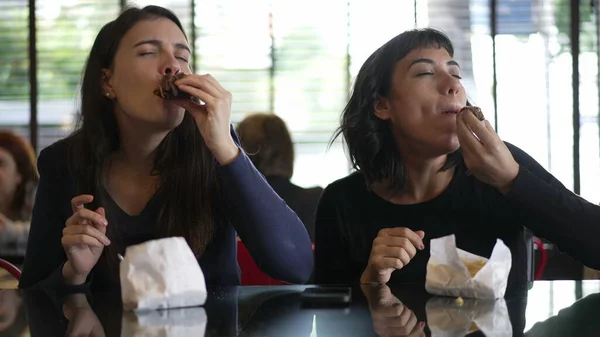 Two women eating burgers for lunch. Female girlfriends taking a bite of hamburgers together at the same time