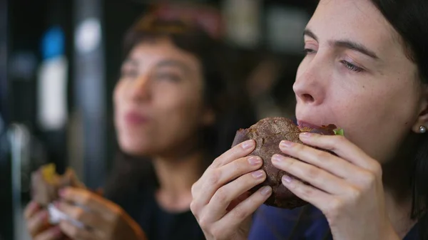 Two people eating burgers. Young women taking a bite of cheeseburgers. Female friends eating fast food lunch