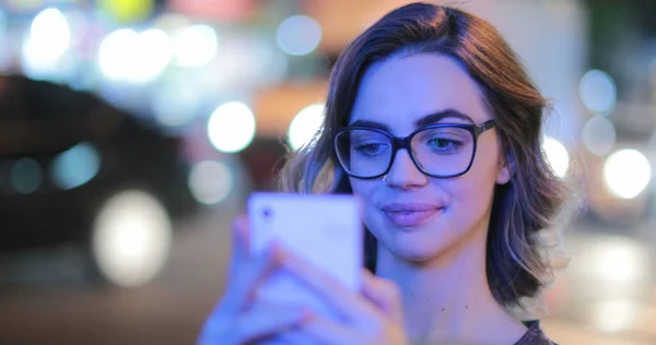 Girl wearing glasses checking cellphone at night in city atmosphere