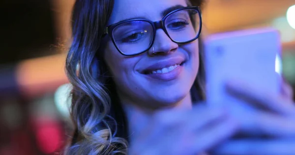 Girl wearing glasses checking cellphone at night in city atmosphere. Young woman smiling holding smartphone in the evening