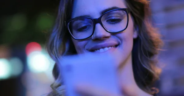 Girl wearing glasses checking cellphone at night in city atmosphere. Young woman smiling holding smartphone in the evening