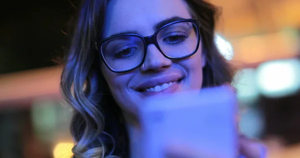 Girl wearing glasses checking cellphone at night in city atmosphere. Young woman smiling holding smartphone in the evening