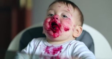Adorable baby covered with red sauce. Feeding messy infant on high-chair