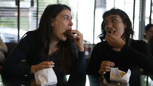 Two girlfriends eating burgers at restaurant for lunch. Female friends hanging out while eating fast food