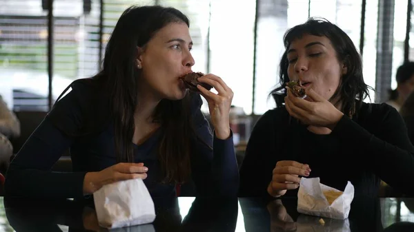 Two girlfriends eating burgers at restaurant for lunch. Female friends hanging out while eating fast food