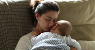 Mother caring for sleeping baby on sofa at home. Casual candid and authentic mom loving her infant son