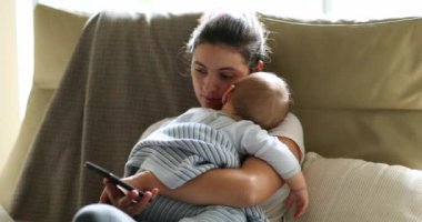Mother checking cellphone device while holding sleeping baby infant on sofa