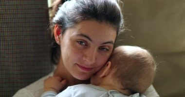 Mother holding baby smiling to camera. Real life authentic casual mom looking to camera holding sleeping infant