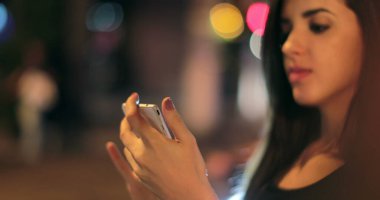 Young hispanic brunette woman checking her cellphone at night. Latina girl holding her smartphone in the evening next to traffic lights