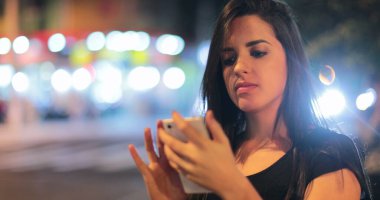 Young hispanic brunette woman checking her cellphone at night. Latina girl holding her smartphone in the evening next to traffic lights