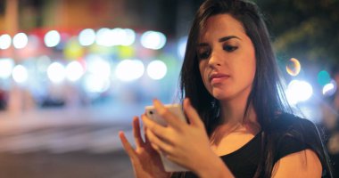 Young hispanic brunette woman checking her cellphone at night. Latina girl holding her smartphone in the evening next to traffic lights