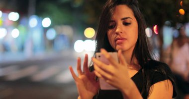 Young hispanic brunette woman checking her cellphone at night. Latina girl holding her smartphone in the evening next to traffic lights