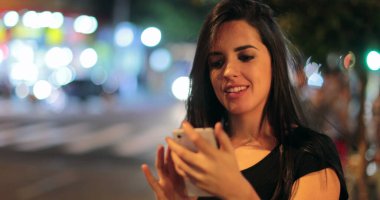 Young hispanic brunette woman checking her cellphone at night. Latina girl holding her smartphone in the evening next to traffic lights