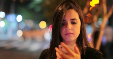 Young hispanic brunette woman checking her cellphone at night. Latina girl holding her smartphone in the evening next to traffic lights