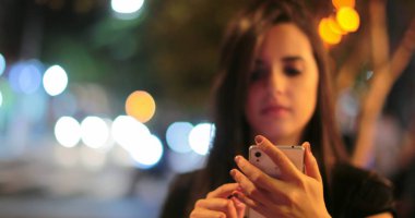 Young hispanic brunette woman checking her cellphone at night. Latina girl holding her smartphone in the evening next to traffic lights