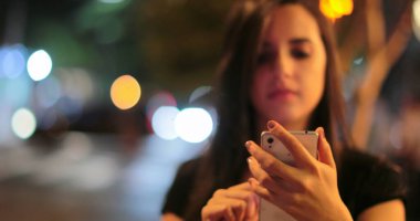 Young hispanic brunette woman checking her cellphone at night. Latina girl holding her smartphone in the evening next to traffic lights