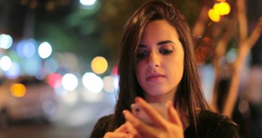 Young hispanic brunette woman checking her cellphone at night. Latina girl holding her smartphone in the evening next to traffic lights