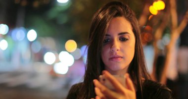 Young hispanic brunette woman checking her cellphone at night. Latina girl holding her smartphone in the evening next to traffic lights