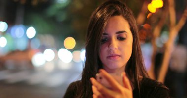 Young hispanic brunette woman checking her cellphone at night. Latina girl holding her smartphone in the evening next to traffic lights