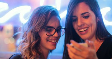 Two girls checking smartphone at night. Young women starring at screen next to neon light