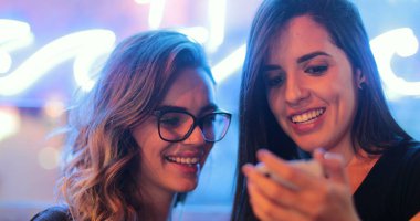 Two girls checking smartphone at night. Young women starring at screen next to neon light
