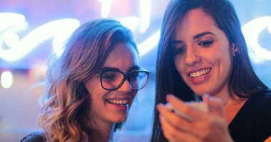 Two girls checking smartphone at night. Young women starring at screen next to neon light