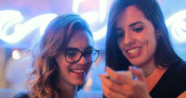 Two girls checking smartphone at night. Young women starring at screen next to neon light