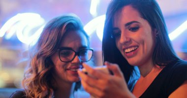 Two girls checking smartphone at night. Young women starring at screen next to neon light
