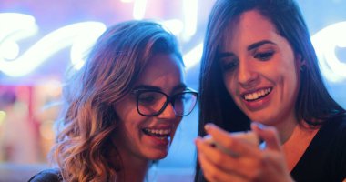 Two girls checking smartphone at night. Young women starring at screen next to neon light