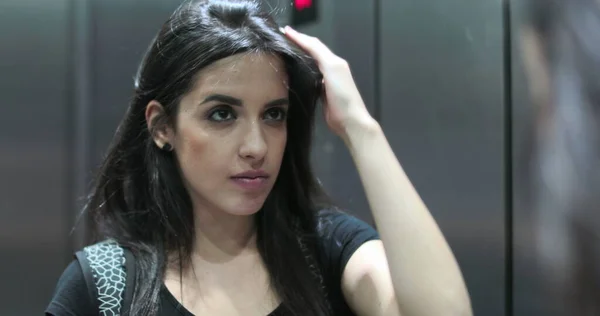 Young hispanic latina woman checking herself in the elevator mirror. Re-arranging hair starring in front of her reflection