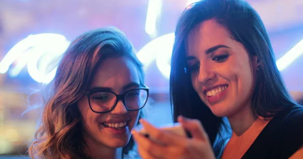 Two girls checking smartphone at night. Young women starring at screen next to neon light