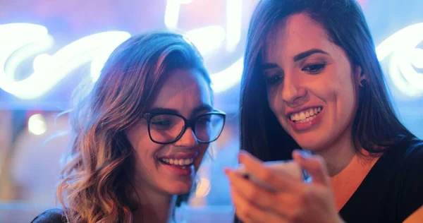 Two girls checking smartphone at night. Young women starring at screen next to neon light