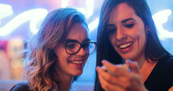 Two girls checking smartphone at night. Young women starring at screen next to neon light