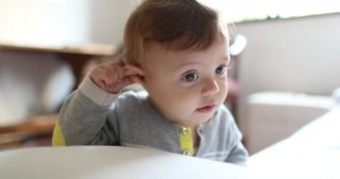 Adorable baby playing with ear. Cute toddler child holding into couch sofa touching ear