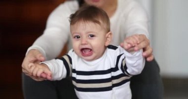 Casual family moment of baby toddler with mom looking to camera smiling, learning to stand up