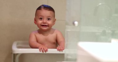 Bathing cute baby infant boy. Expressive toddler child holding into side of bathtub smiling being adorable